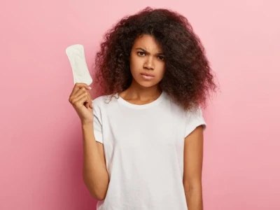 Uma mulher veste uma camiseta branca e segura um absorvente higiênico. Ela tem uma expressão de frustração e confusão, com a sobrancelha franzida, em frente a um fundo rosa.