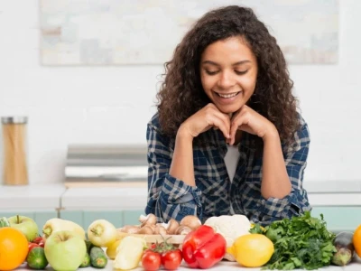 Jovem mulher sorrindo em uma cozinha, olhando para uma mesa repleta de frutas e vegetais coloridos, demonstrando a importância da variedade em uma alimentação saudável.