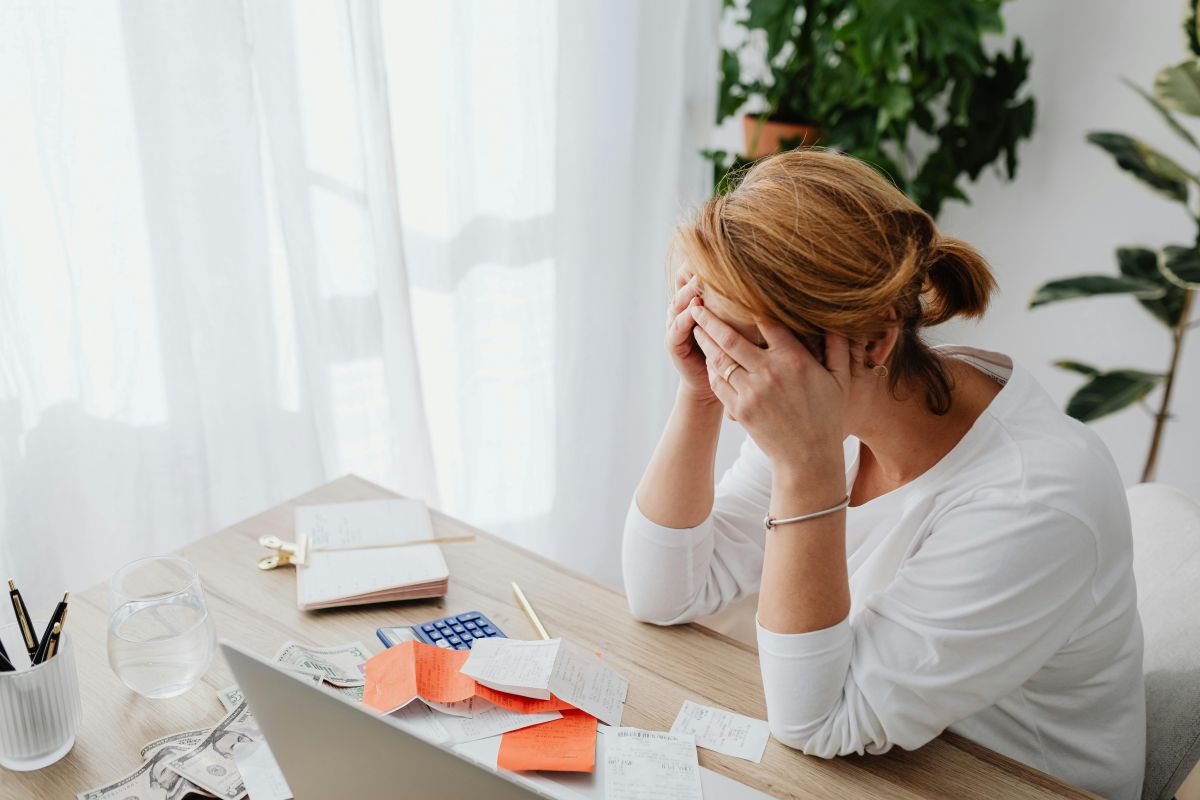 Uma mulher, com as mãos no rosto, parece estar estressada ou sobrecarregada em sua mesa de trabalho com papéis e um laptop, simbolizando o esgotamento que multivitaminicos podem ajudar a combater.