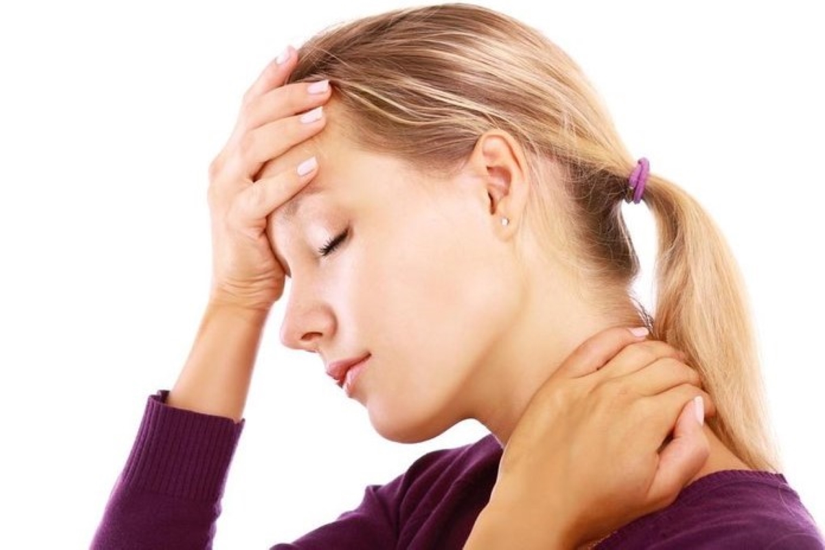 Mulher com expressão de desconforto, levando a mão à cabeça e ao pescoço, simbolizando sintomas comuns da TPM como dor de cabeça, irritação e tensão muscular. Fundo branco e roupa roxa.