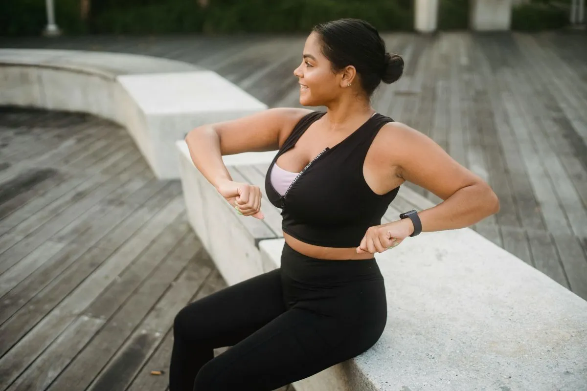 Uma mulher sorridente com roupas de ginástica pretas sentada em um banco de concreto ao ar livre, se preparando para o exercício com energia.