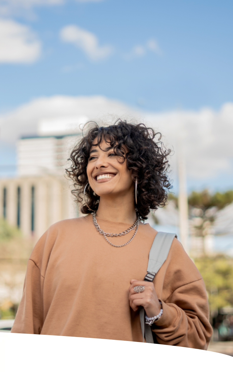 Jovem mulher caminhando em ambiente urbano, sorridente e carregando bolsa.