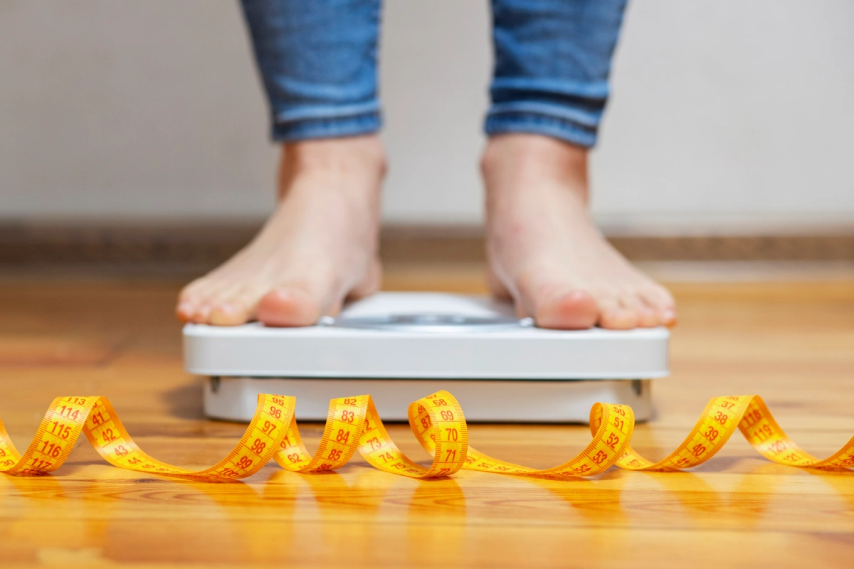 Pés de uma mulher em cima de balança de piso, com uma fita métrica ao lado, checando sua pesagem