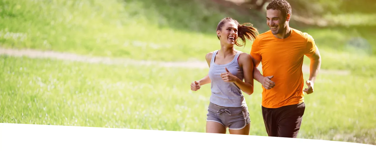 Casal correndo feliz por uma trilha de parque, durante uma tarde ensolarada.
