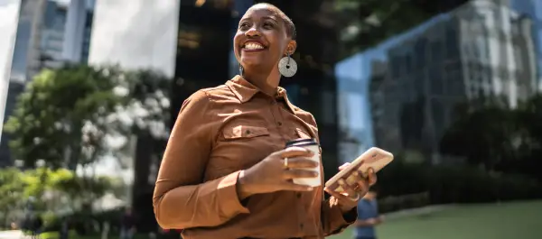 Mulher sorridente, em famosa avenida de São Paulo, segurando um café com uma mão e um smartphone com outra.