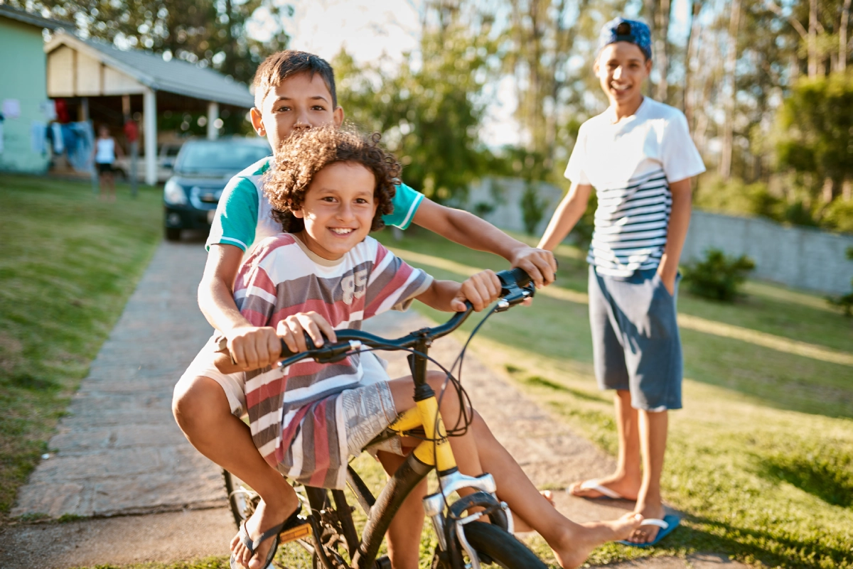 Dois meninos se divertem andando em uma mesma bicicleta, em uma tarde ensolarada, no quintal, sob a supervisão de um adulto.