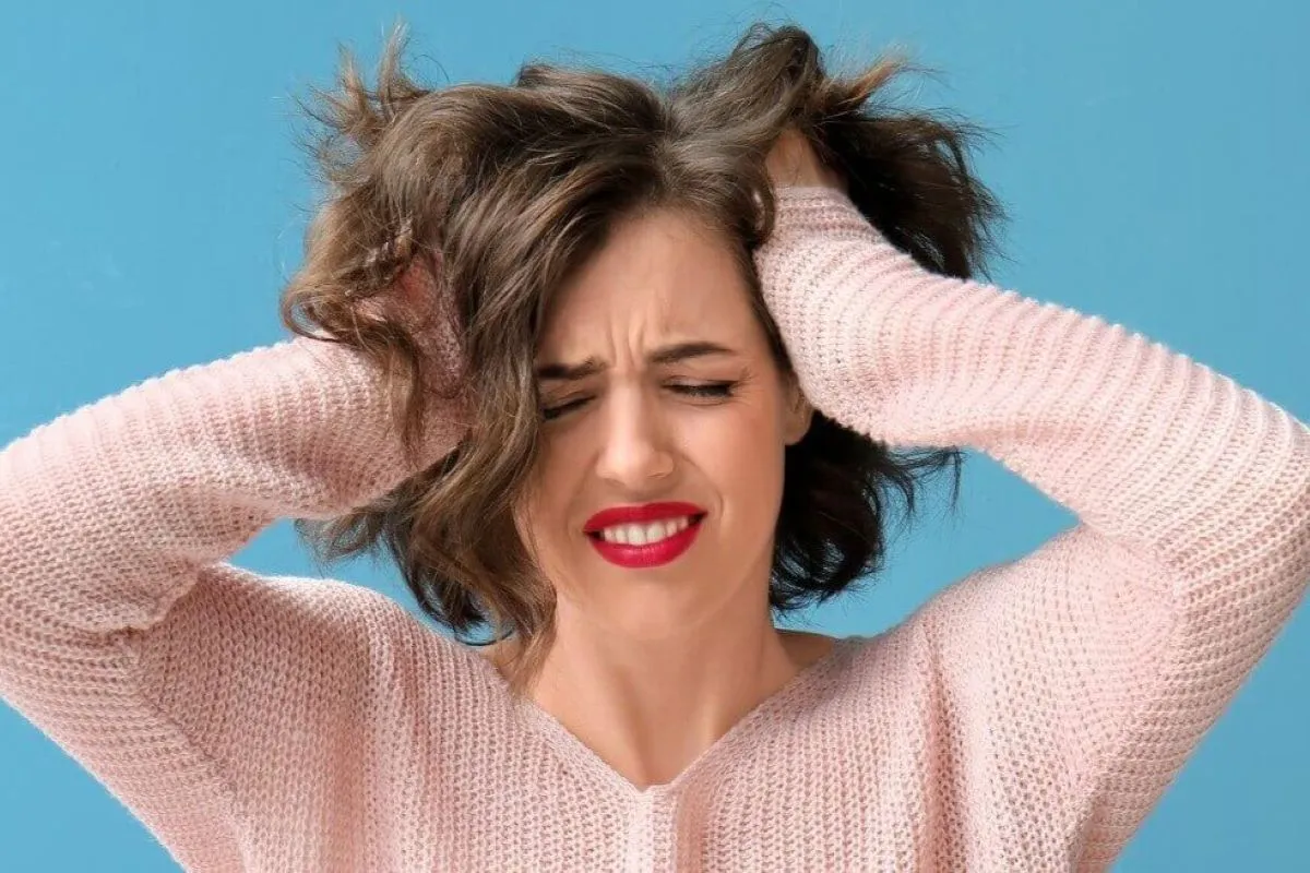 Mulher de cabelos cacheados e blusa rosa, com os olhos fechados e dentes cerrados, segurando a cabeça com as mãos em um gesto de estresse, sobre fundo azul.