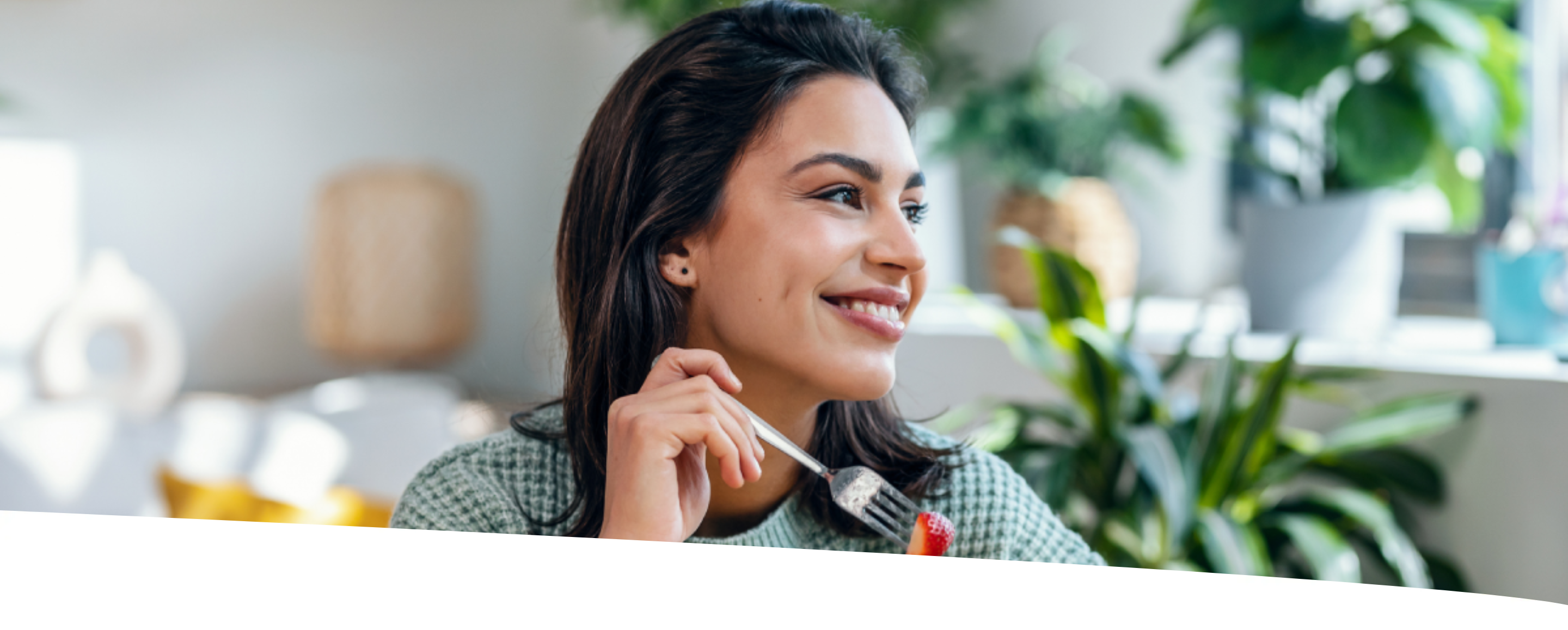 Mulher sorridente comendo um prato de salada com morangos, em uma casa com plantas, olhando para a janela, num dia de sol.