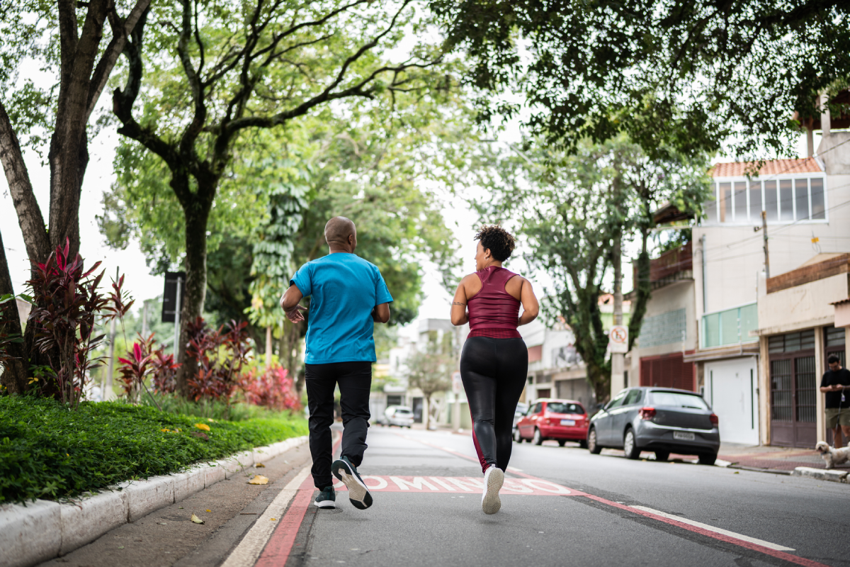 Casal fazendo jogging na rua, durante o dia, sorrindo um para o outro.