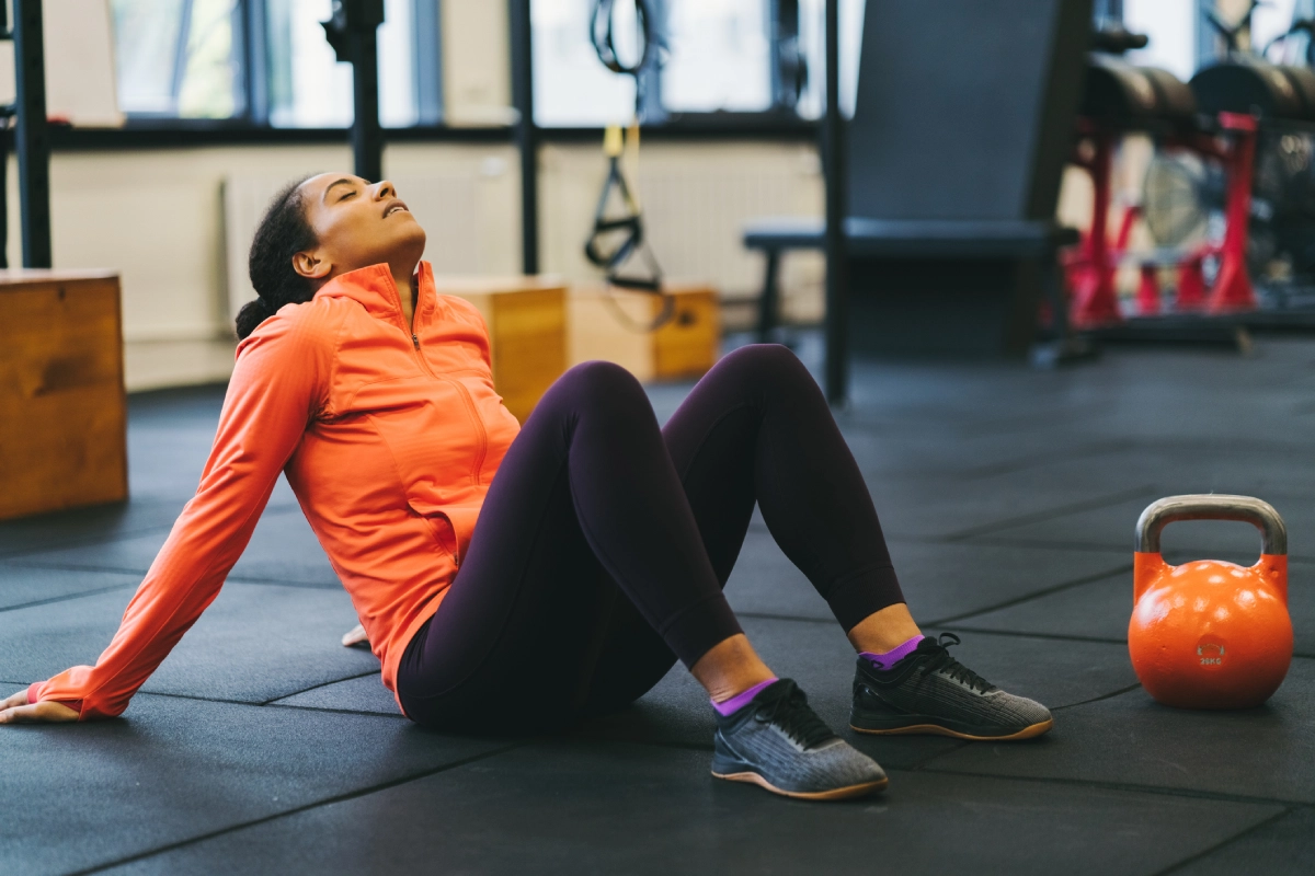 Mulher na academia, sentada no chão com os pés no chão e joelhos flexionados, jogando a cabeça para trás e apoiada nas mãos, descansando entre séries.