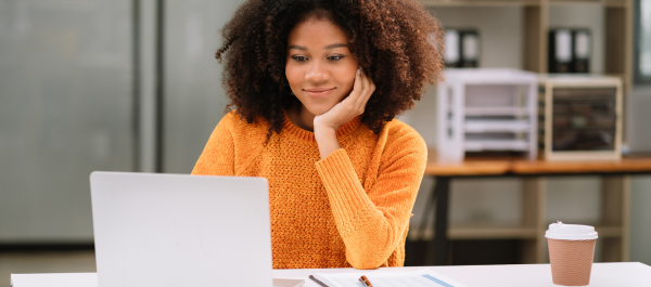 Garota sentada em mesa de escritório, com sorriso no rosto, mão esquerda apoiando o queixo e a outra usando um laptop.