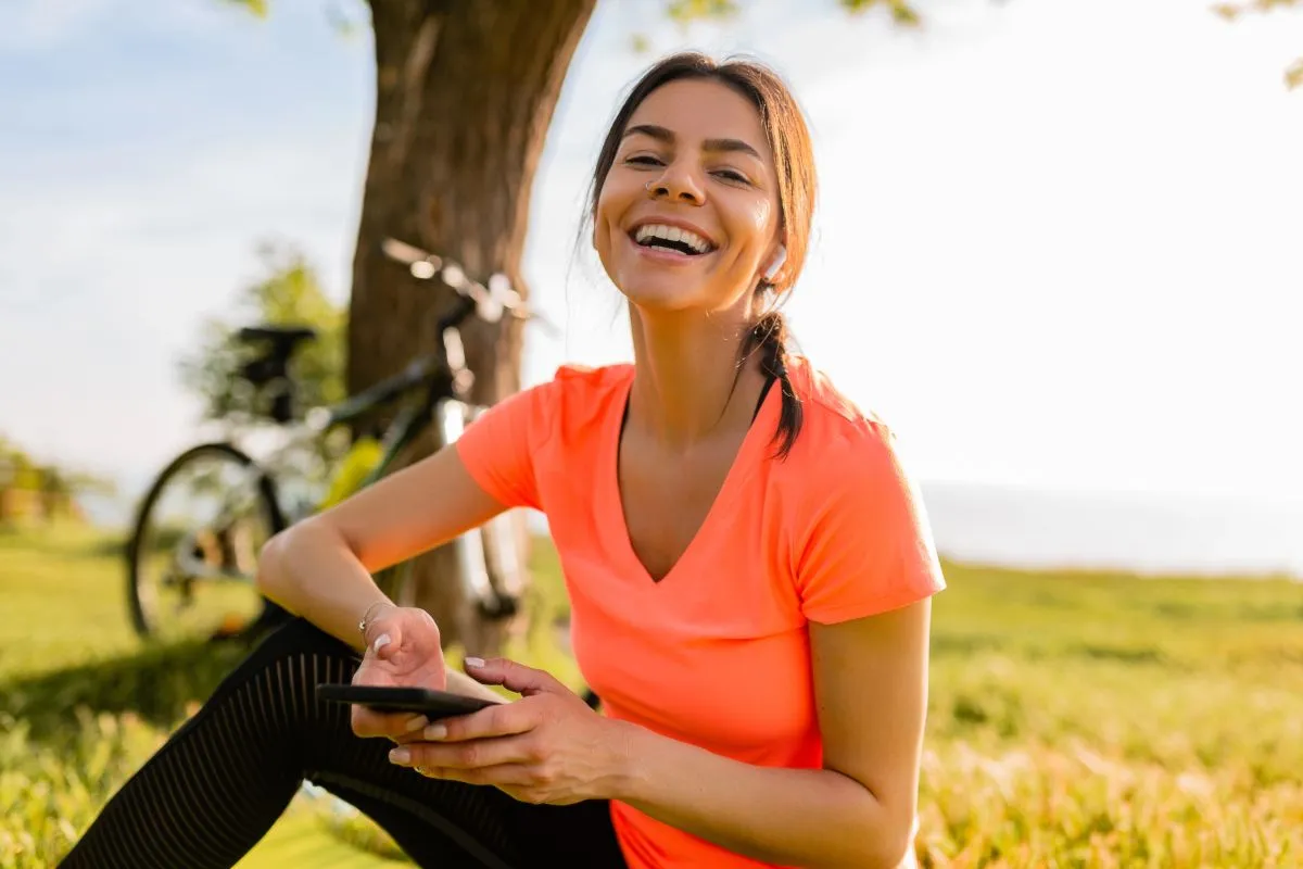Mulher sorridente sentada na grama de um parque, usando fones de ouvido e segurando um celular, com uma árvore e uma bicicleta ao fundo durante o pôr do sol.