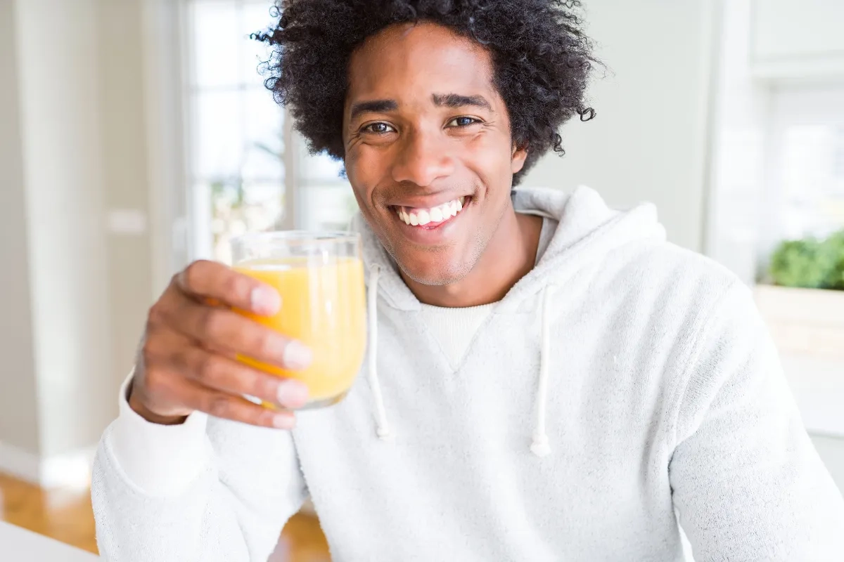 Homem sorridente tomando um suco de laranja e olhando para a câmera (a foto parece uma selfie).