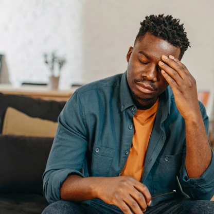 Homem sentado no sofá com expressão cansada e a mão na cabeça, ilustrando sinais de estresse no dia a dia.