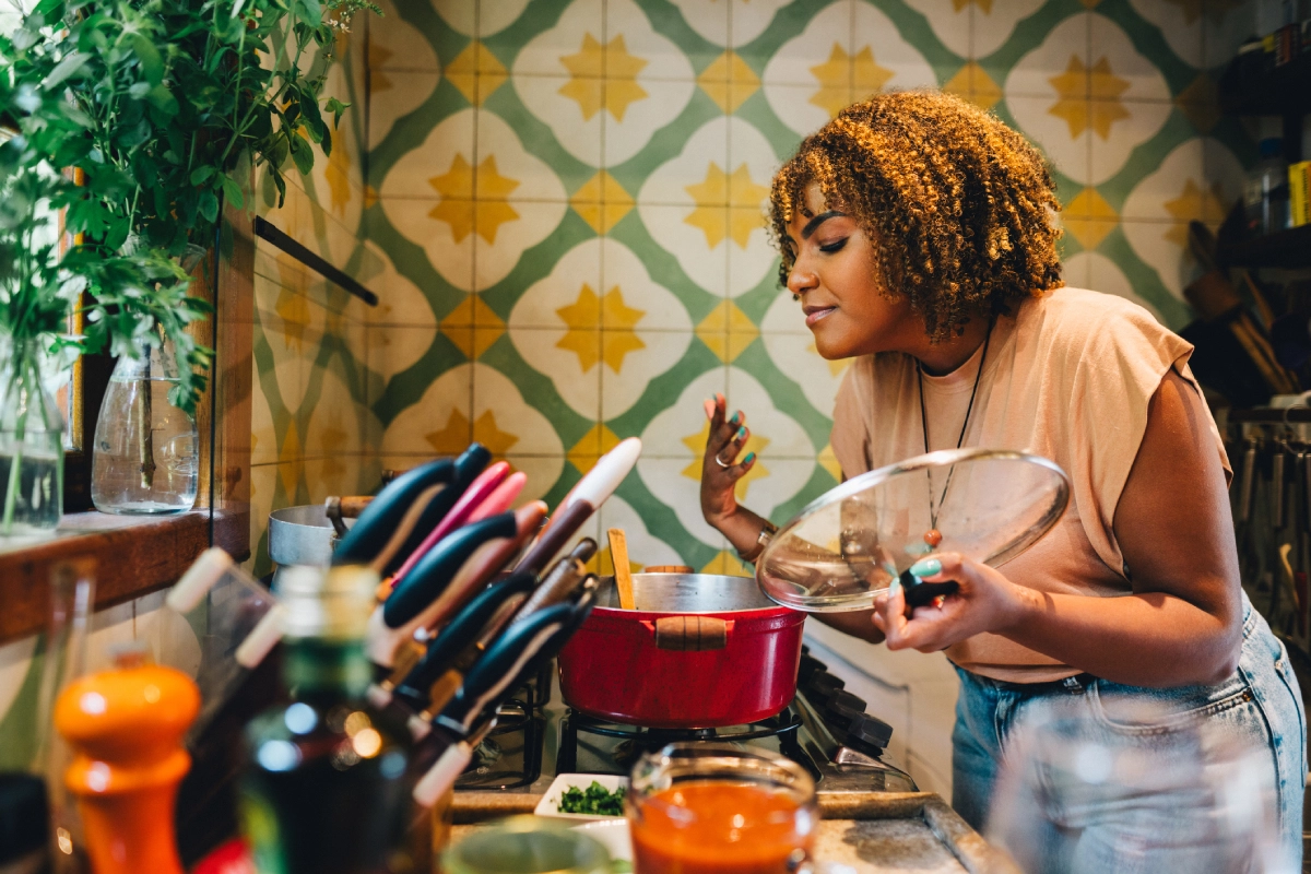 Mulher em cozinha aconchegante e vintage abre uma panela para sentir o aroma do que está sendo preparado.