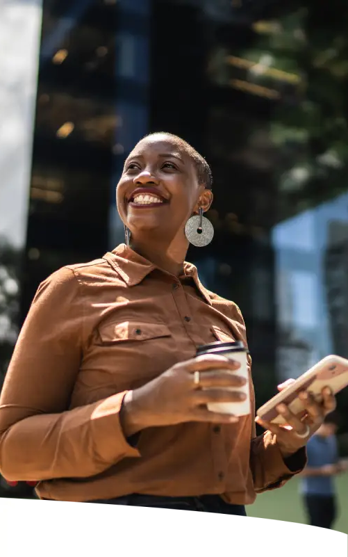 Mulher sorridente, em famosa avenida de São Paulo, segurando um café com uma mão e um smartphone com outra.