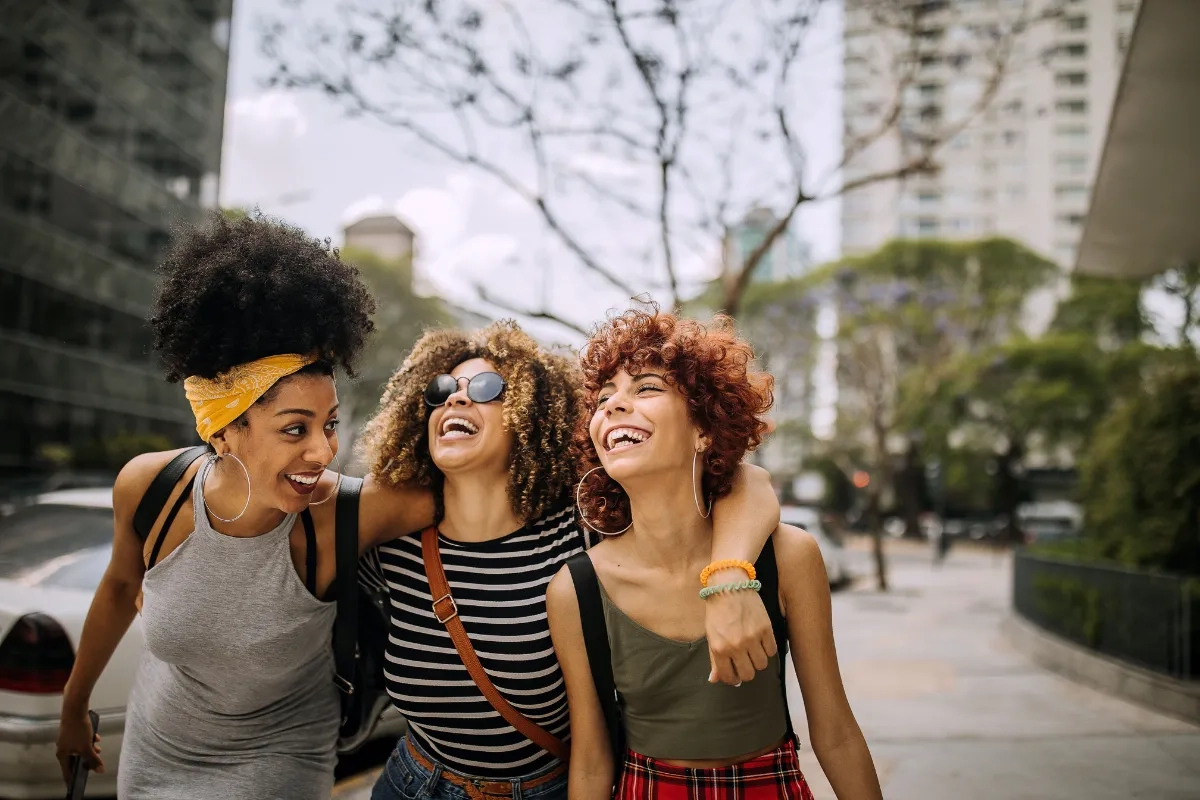 Três amigas caminhando abraçadas e sorridentes em um ambiente urbano.