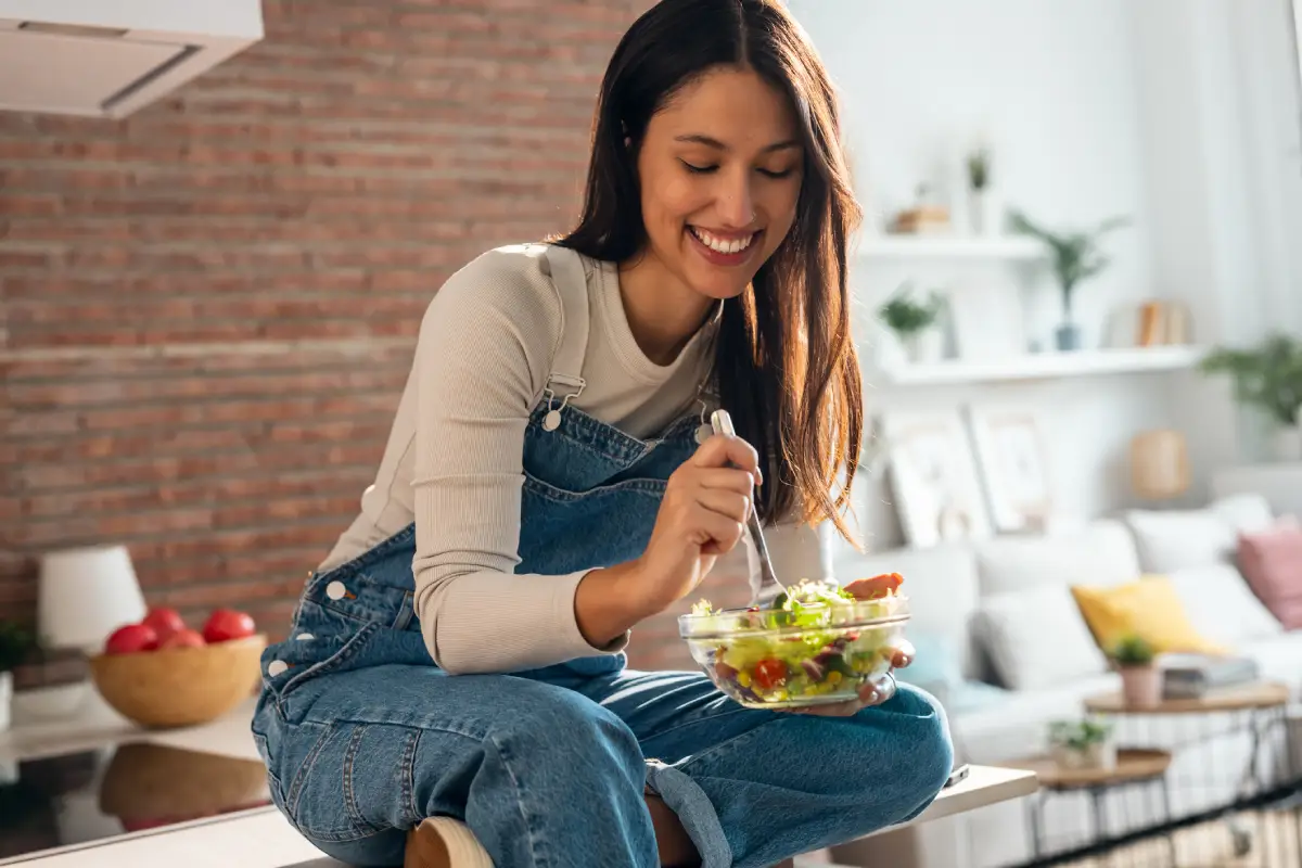 Mulher sentada sobre a ilha da cozinha, sorrindo enquanto come uma salada.