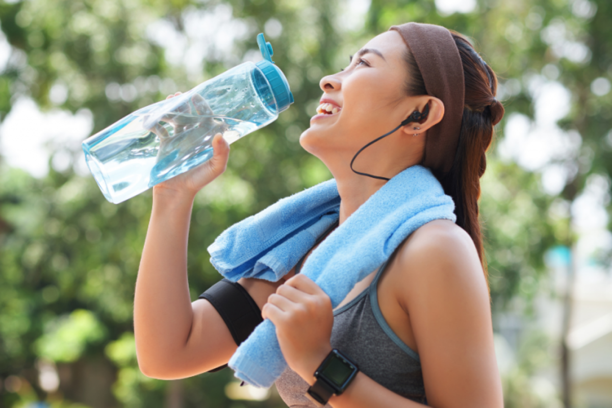 Mulher sorridente bebendo água após a prática de exercícios físicos ao ar livre. Ela usa roupas esportivas e fones de ouvido, representando um estilo de vida saudável com hidratação, energia e bem-estar.