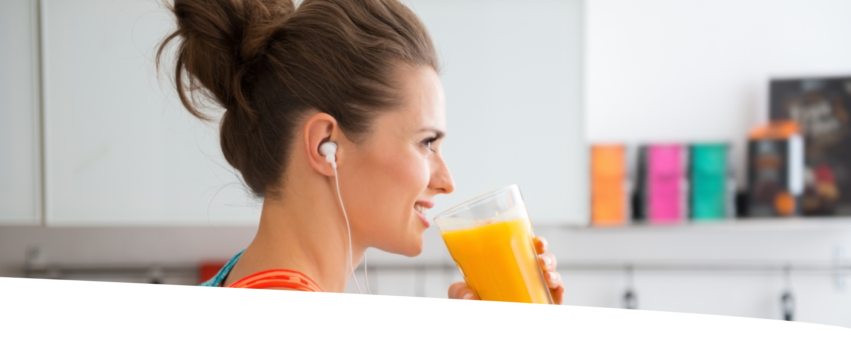Mulher com coque e roupas leves, usando fones de ouvido, está na cozinha tomando um suco de laranja.