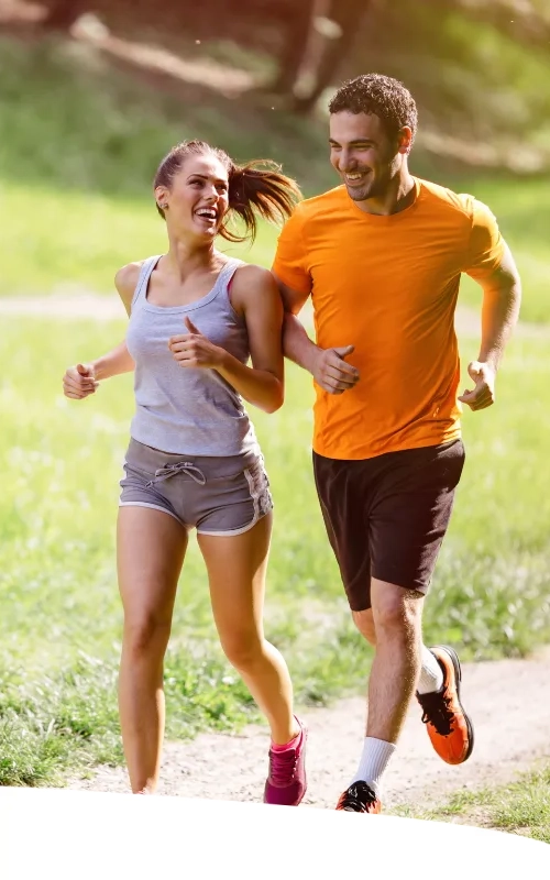 Casal correndo feliz por uma trilha de parque, durante uma tarde ensolarada.