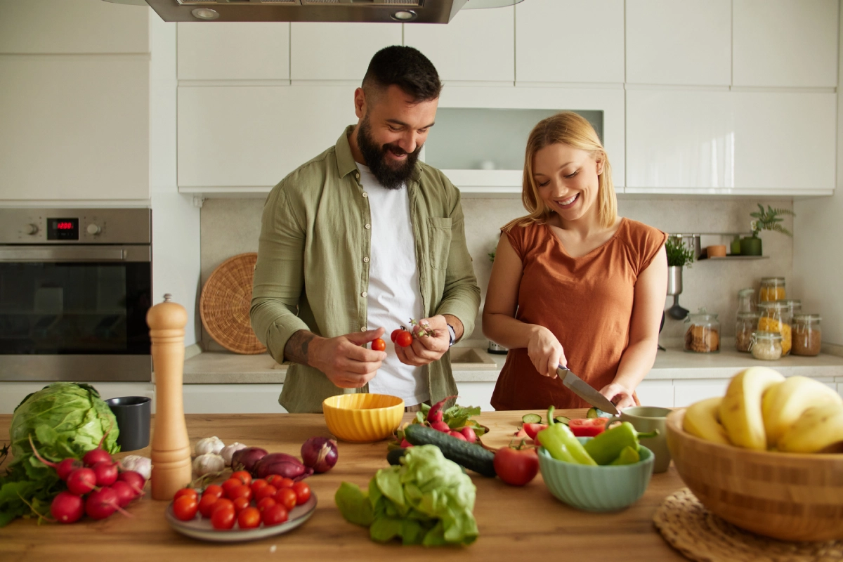 Casal feliz preparando alimentos na cozinha. Sobre a bancada, há uma série de frutas e verduras.