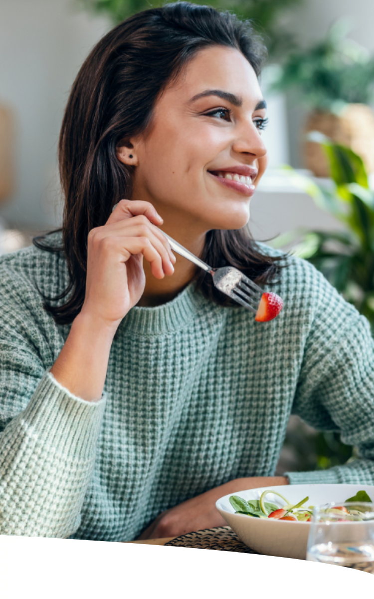 Mulher sorridente comendo um prato de salada com morangos, em uma casa com plantas, olhando para a janela, num dia de sol.