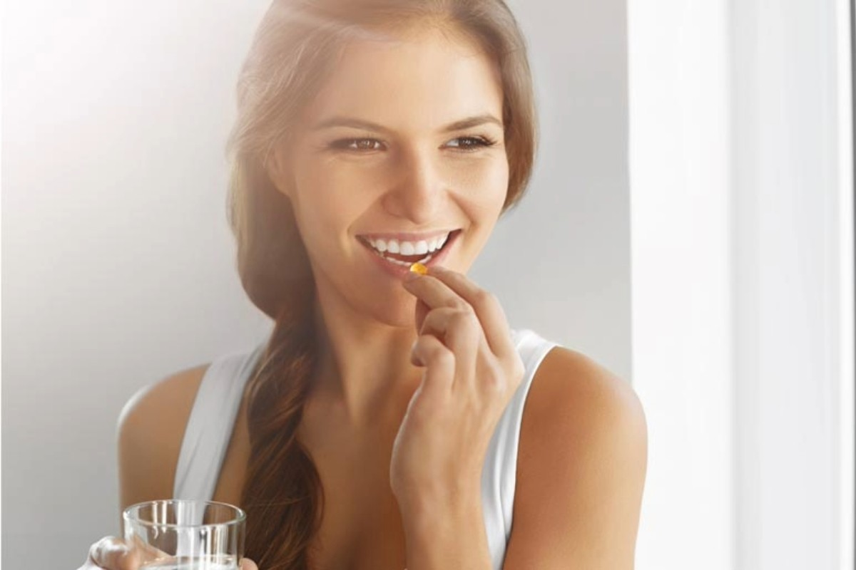 Mulher sorridente segurando um copo de água em uma mão e uma cápsula ou comprimido na outra, prestes a tomá-lo. Ela está em um ambiente iluminado, próximo a uma janela, transmitindo uma sensação de saúde e bem-estar.
