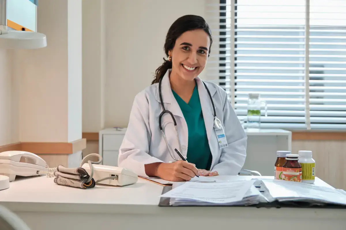 Nutróloga em seu escritório, segurando caneta e sorrindo para a foto.