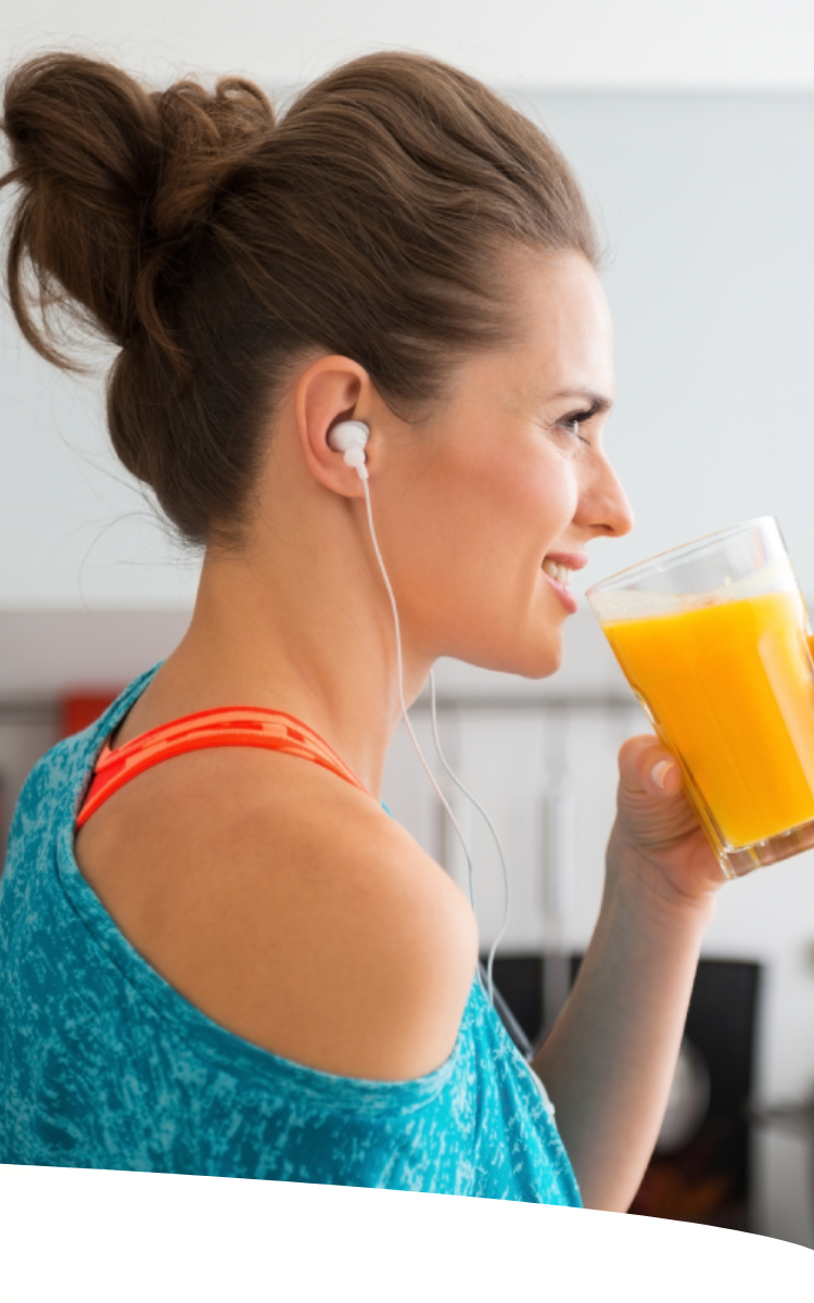 Mulher com coque e roupas leves, usando fones de ouvido, está na cozinha tomando um suco de laranja.