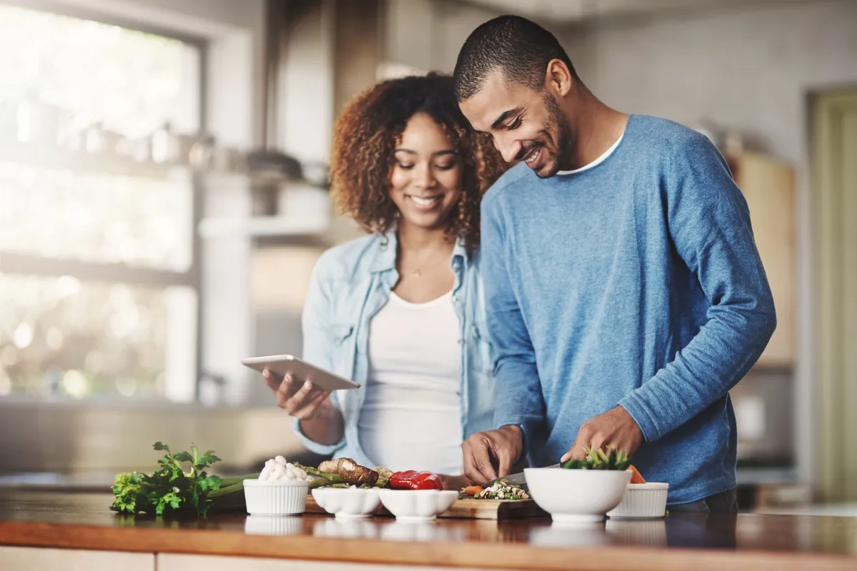Casal está preparando uma refeição juntos na cozinha, seguindo uma receita que a mulher parece ler no celular.