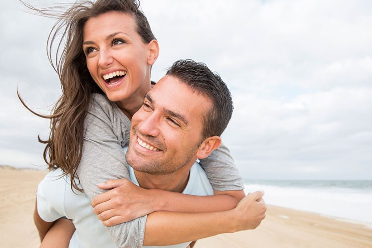 Casal sorrindo na praia olhando para o lado. Homem segura mulher nas costas. Ao fundo, é possível ver areia e água da praia.