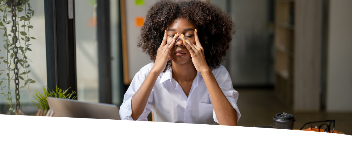Uma mulher com as mãos no rosto, sentada em sua estação de trabalho, parece estar exausta.