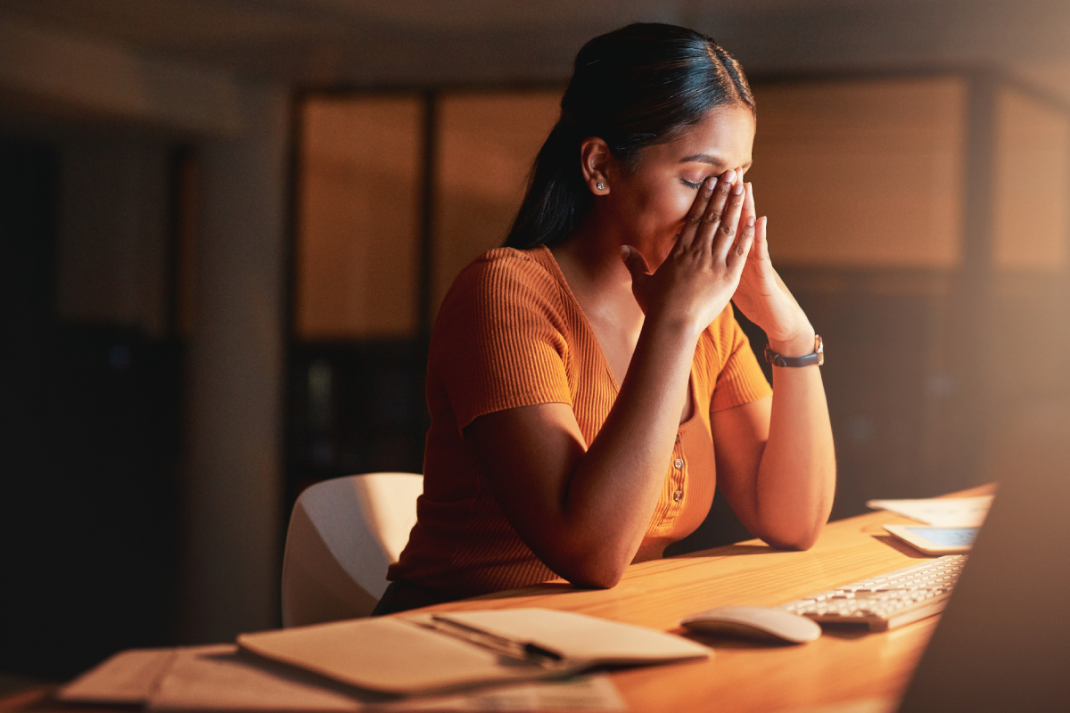 Mulher estressada trabalhando à noite no escritório, com as mãos no rosto, mostrando sinais de cansaço mental e exaustão profissional. Ambiente com iluminação suave, computador e caderno sobre a mesa.