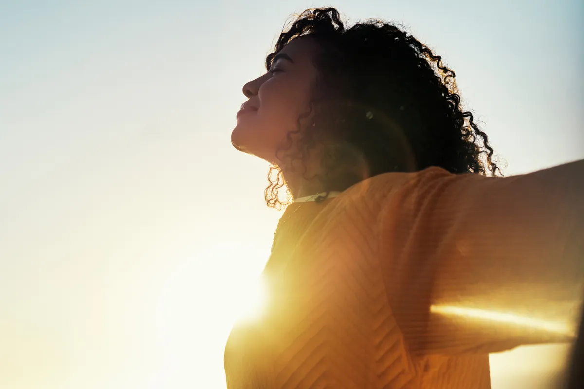Mulher de braços abertos e cabeça erguida - na foto, o Sol aparece brilhante na altura do seu peito.