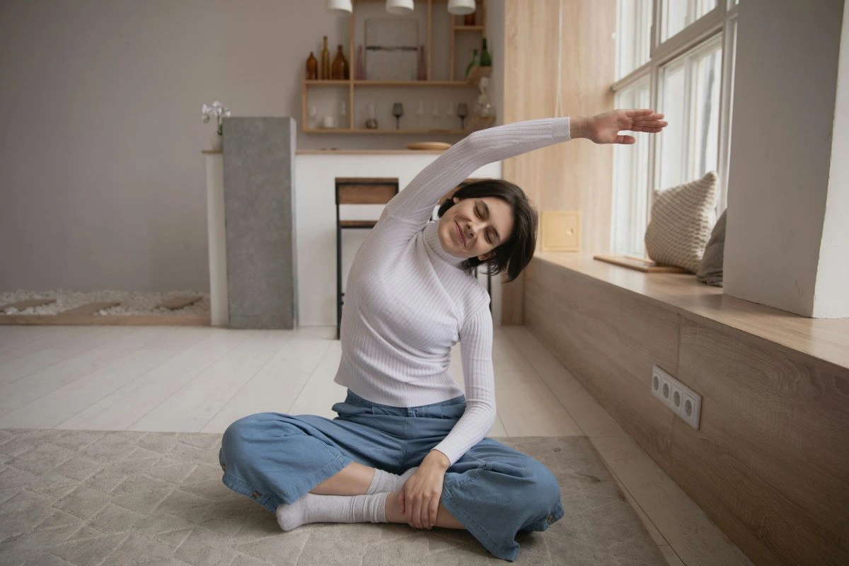 Uma mulher praticando yoga, sentada em posição de lótus e realizando um alongamento lateral do tronco, em um ambiente interno bem iluminado.