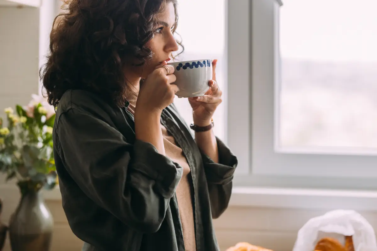 Mulher levando xícara de café à boca com as duas mãos, enquanto olha pensativa pela janela.