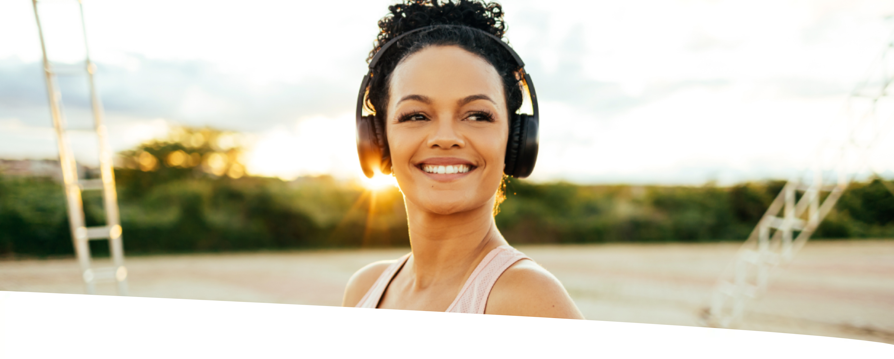 Mulher sorridente, com fone de ouvido, porta-celular no braço e garrafa de chá em mãos, no parque em uma tarde ensolarada.