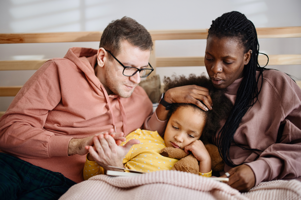 Pai e mãe estão na cama, cuidando da filha que parece doente, e está na cama lendo um livro.