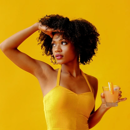 Mulher segurando um copo de suco enquanto observa o ambiente, em um cenário amarelo que remete à vitamina C e aos benefícios para a imunidade.