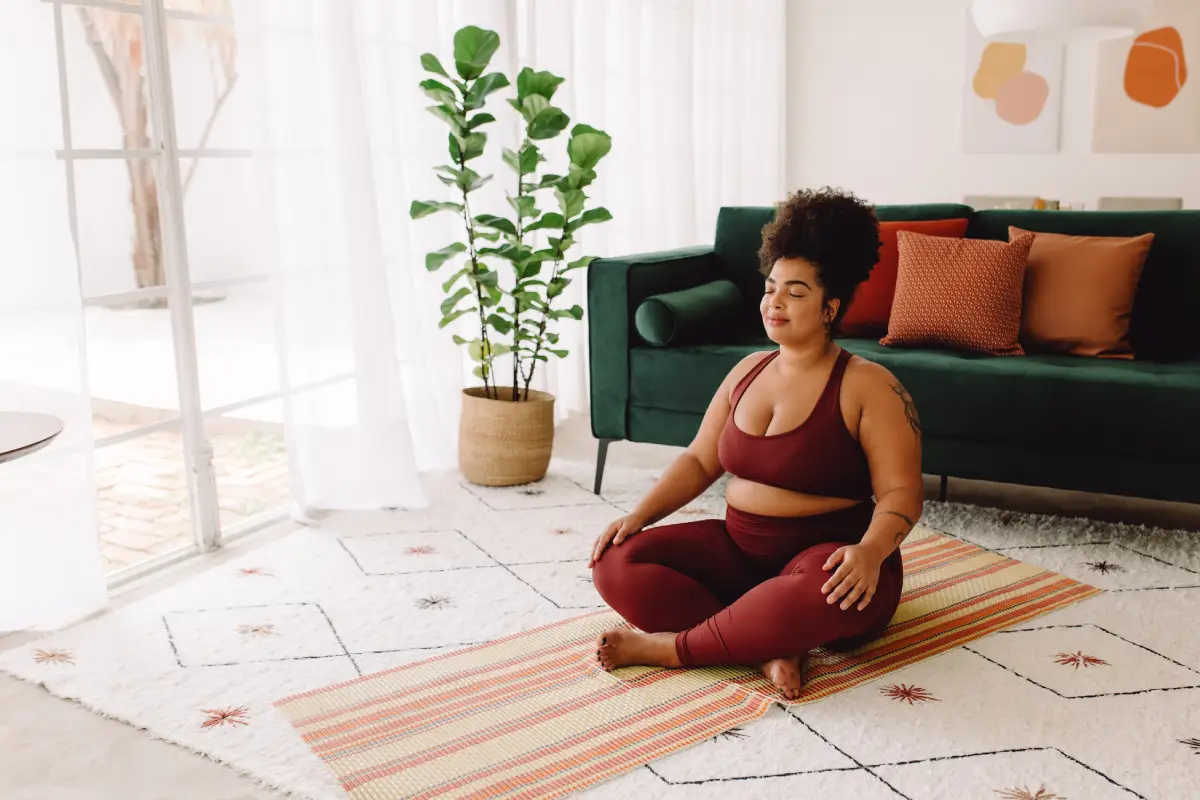 Mulher sentada no meio da sala, sobre tapete de ioga, em posição de meditação.