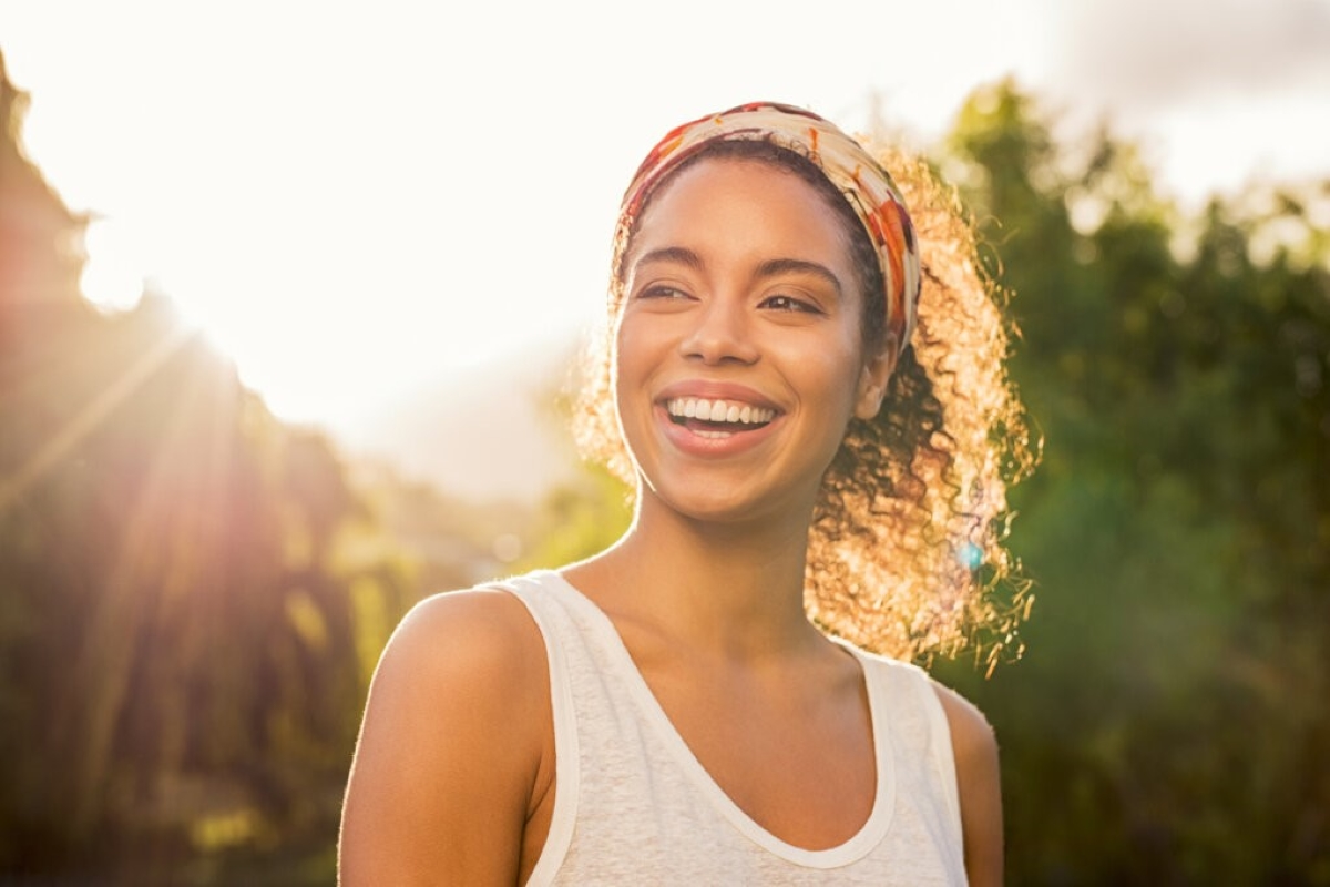 Mulher sorridente ao ar livre, com aparência de vitalidade e bem-estar. Ela veste uma camiseta clara e está em um ambiente iluminado pelo sol, representando os benefícios da vitamina C para a energia e imunidade.