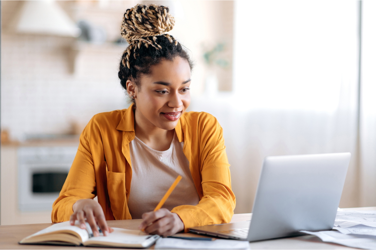 Mulher estudando em laptop enquanto faz anotações em um caderno.