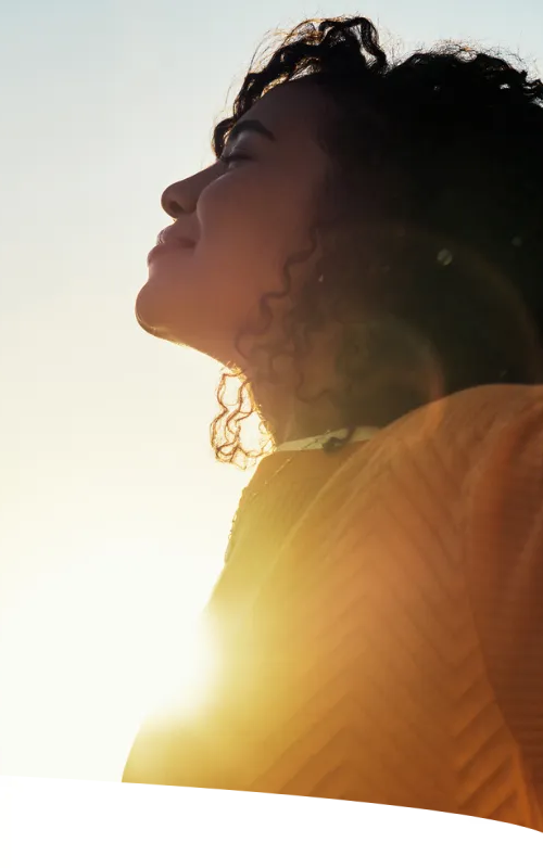 Mulher de braços abertos e cabeça erguida - na foto, o Sol aparece brilhante na altura do seu peito.