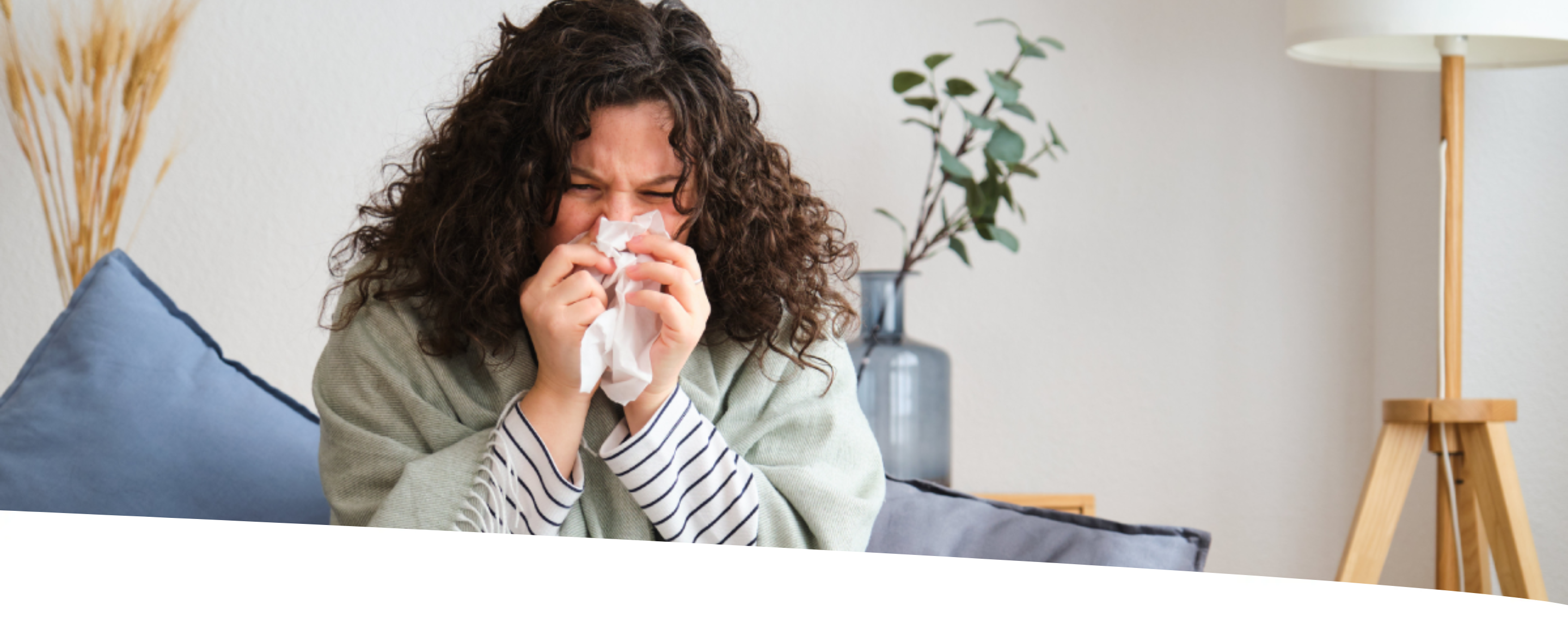 Mulher sentada no sofá, muito agasalhada, assoando o nariz em um lenço de papel.