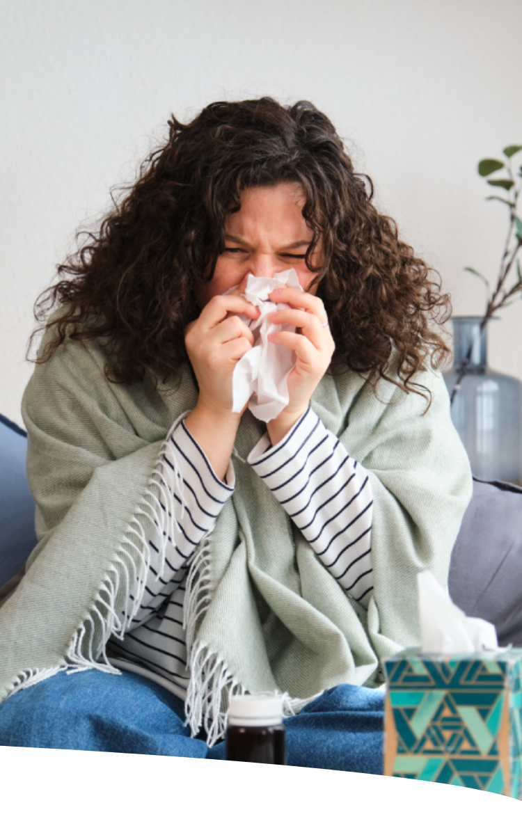 Mulher sentada no sofá, muito agasalhada, assoando o nariz em um lenço de papel.