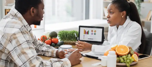 Homem conversando com sua nutricionista. Sobre a mesa, vemos alimentos frescos e suplementos.