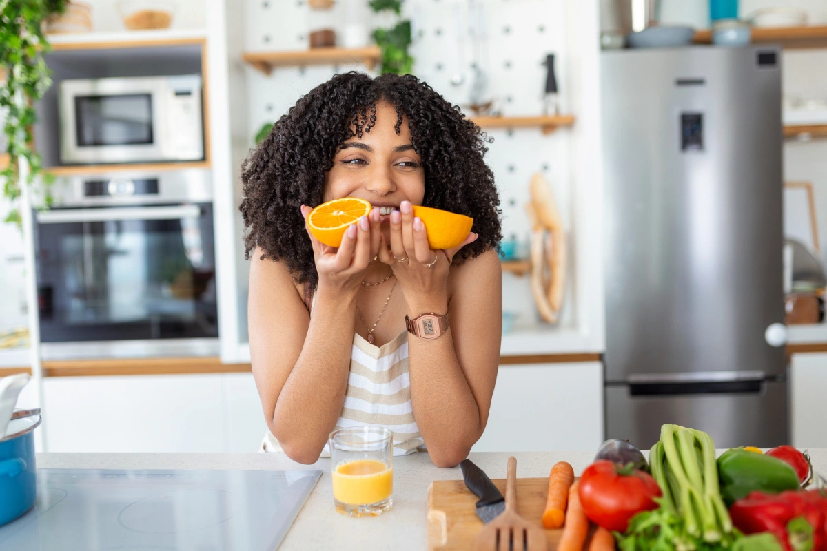 Mulher sorridente na cozinha segura duas metades de laranja próximo ao rosto