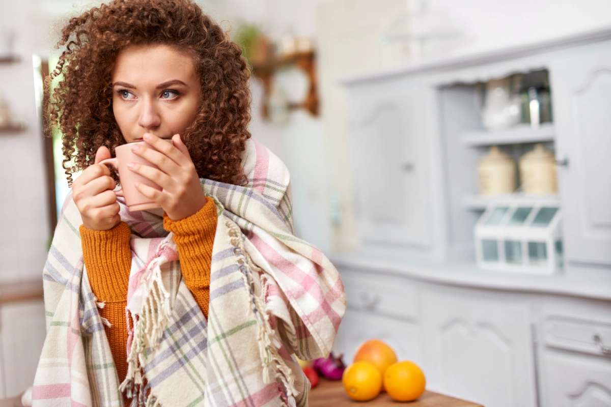 Mulher está em sua cozinha, coberta com uma manta confortável, levando à boca uma caneca de chá com as duas mãos.