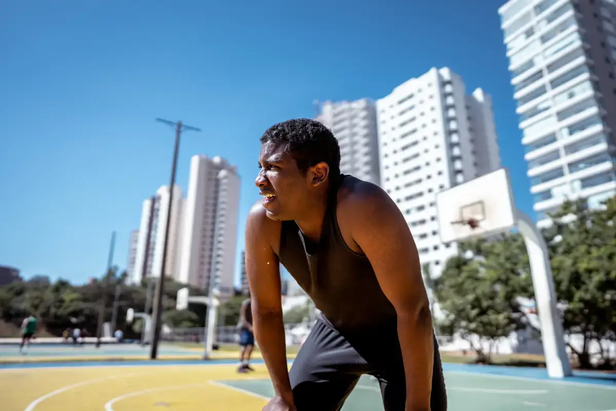 Homem está em meio a uma quadra de basquete, em dia ensolarado, apoiando as mãos nos joelhos para descansar.